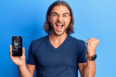 Kırık akıllı telefonu tutan genç yakışıklı adam kırık ekranı gösteriyor gururla bağırıyor, zaferi kutluyor ve başarıyı kollarını kaldırarak kutluyor. 
