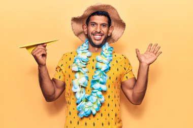 Yaz şapkası takan genç bir latin ve elinde kağıt uçakla zaferi kutlayan Hawaii zambağı tutan mutlu bir gülümseme ve ellerini kaldırarak kazanan bir ifade. 