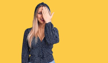 Genç, güzel, sarışın bir kadın gündelik kıyafetler giyiyor. Bir gözünü eliyle kapatıyor. Yüzünde kendine güvenen bir gülümseme ve sürpriz bir duygu var.. 