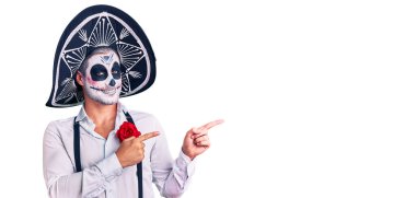 Arka planda ölü kostümü giymiş bir adam gülümsüyor ve kameraya bakıyor iki eliyle ve parmaklarıyla kenarı işaret ediyor.. 