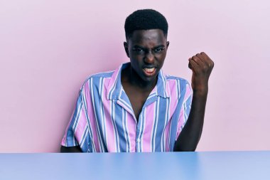Genç, Afro-Amerikan bir adam, gündelik giysiler giymiş, kızgın ve kızgın bir şekilde masaya oturmuş öfkeyle bağırırken yumruğunu kaldırıyor. öfke ve agresif konsept. 