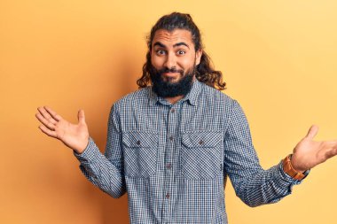 Gündelik giysiler giyen genç bir Arap, elleri ve kolları havada, kafası karışık bir ifade takınıyor. şüphe kavramı. 