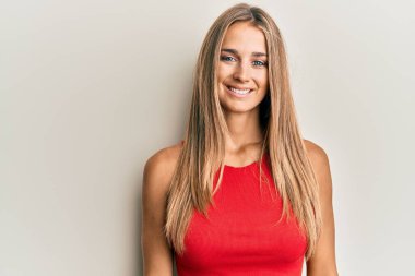 Gündelik kıyafetler giyen genç sarışın kadın pozitif ve mutlu görünüyor. Kendine güvenen bir gülümsemeyle ayakta duruyor ve dişlerini gösteriyor. 