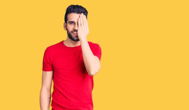 Sakallı, sıradan tişörtler giyen, bir gözünü eliyle kapatan, yüzünde kendine güvenen bir gülümseme ve sürpriz duygular olan genç yakışıklı bir adam.. 