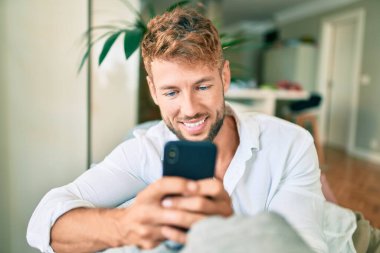 Yakışıklı beyaz adam mutlu gülümsüyor. Evdeki kanepede otururken akıllı telefon kullanıyor.