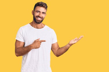 Latin asıllı genç adam, gündelik beyaz tişört giyip kameraya hayret ediyor ve gülümsüyor.. 