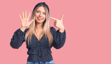 Genç, güzel, sarışın, günlük kıyafetler giyen, yedi numaralı parmaklarıyla işaret eden, kendinden emin ve mutlu bir şekilde gülümseyen bir kadın.. 