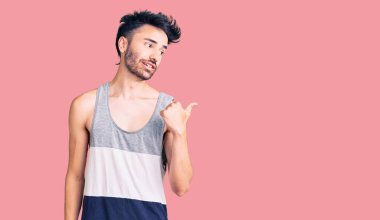 Gündelik kıyafetler giyen, gülümseyen, gülümseyen ve başparmağıyla yan tarafı gösteren İspanyol genç adam.. 