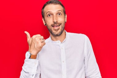 Genç, yakışıklı, günlük kıyafetler giymiş, baş parmağını yana doğru gösteren, ağzı açık gülümseyen bir adam. 