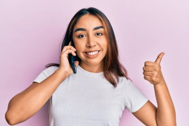 Akıllı telefondan konuşan genç Latin kadın mutlu ve olumlu gülümsüyor, başparmağıyla mükemmel ve onay işareti yapıyor. 