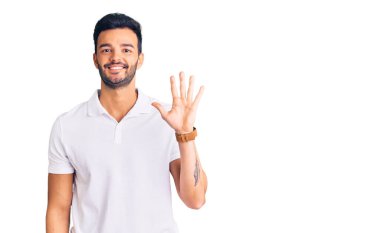 Genç, yakışıklı, günlük kıyafetler giyen, beş numarayı gösterip işaret eden, kendinden emin ve mutlu bir şekilde gülümseyen İspanyol adam.. 