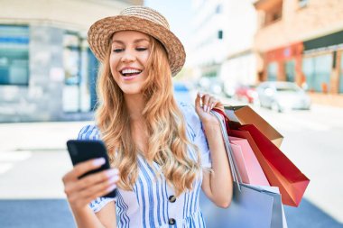 Genç, güzel alışverişçi kadın gülümseyerek markete gidiyor alışveriş torbaları tutuyor, akıllı telefon kullanarak mutlu bir şekilde gülümsüyor.