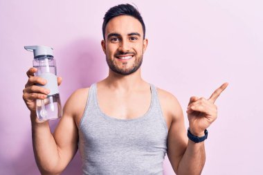 Sakallı genç yakışıklı sporcu kolsuz tişört giyiyor su şişesi içiyor mutlu bir şekilde gülümsüyor.