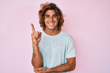 Yüzünde kocaman bir gülümseme olan, gündelik kıyafetler giyen genç İspanyol adam, kameraya el ve parmak ile bakıyor.. 