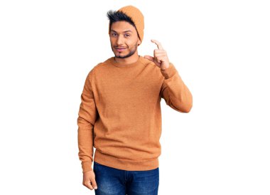 Yakışıklı Latin Amerikalı genç adam kışlık süveter ve yün şapka takıyor gülümsüyor ve kendine güvenen el hareketleriyle küçük beden işareti yapıyor parmaklarıyla ve kamerayla bakıyor. ölçüm kavramı. 