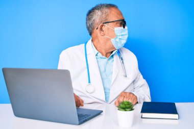 Gri saçlı, doktor üniforması giyen ve tıbbi maske takan, rahat bir görünüm takınan, kendine güvenen gülümseyen yaşlı yakışıklı bir adam.. 