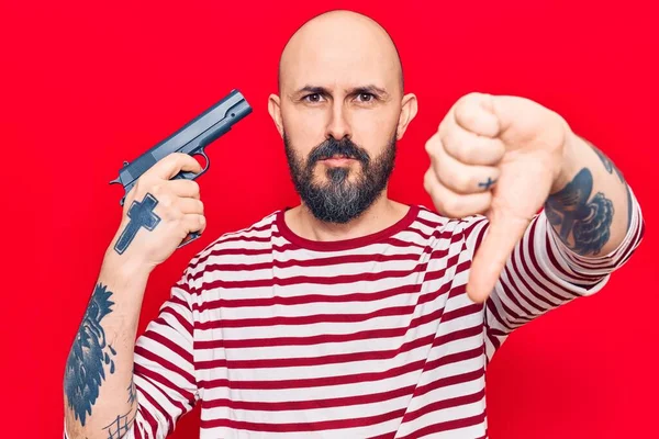 Young handsome man holding gun with angry face, negative sign showing dislike with thumbs down, rejection concept