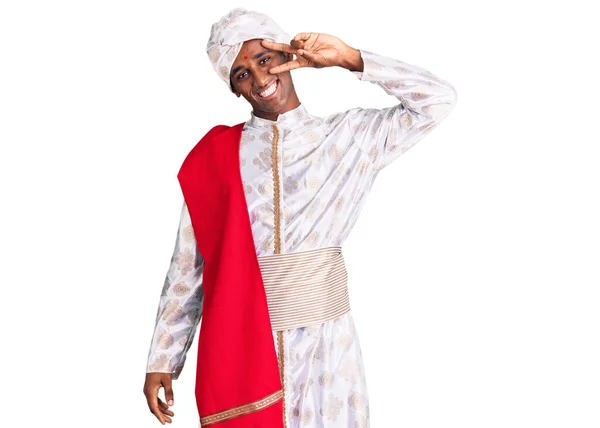 伝統的なシャーワニを身に着けているアフリカのハンサムな男は 顔の上に指で平和のシンボルを行う服を見て 明るく勝利を示す笑みを浮かべて — ストック写真