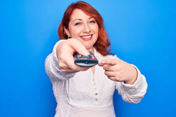 年轻美丽的红头发女人用电视遥控装置换电视频道开心地微笑着用手指头指指点点 — 图库照片