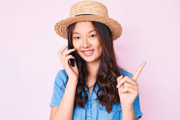 年轻美丽的中国姑娘头戴夏帽 在智能手机上聊天 微笑着 手指手画脚地指向旁边 — 图库照片