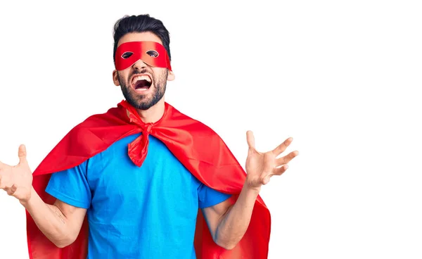 Young handsome man with beard wearing super hero costume crazy and mad shouting and yelling with aggressive expression and arms raised. frustration concept.