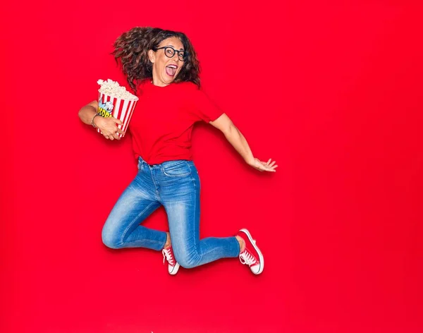 Middle age beautiful hispanic woman surprised with open mouth wearing glasses. Holding bucket of popcorn jumping over isolated red background