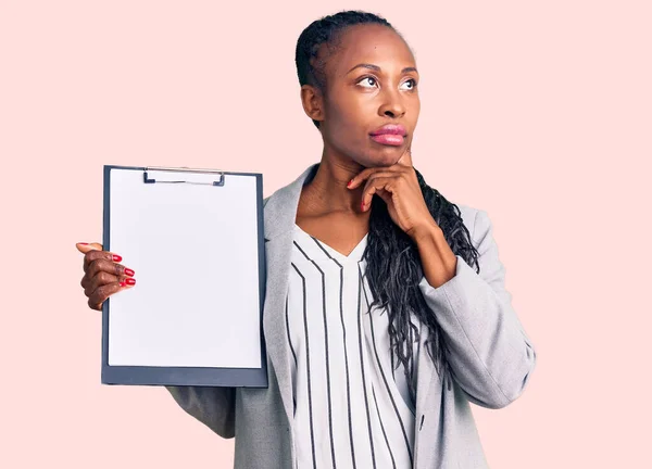 Young african american woman wearing business clothes holding clipboard serious face thinking about question with hand on chin, thoughtful about confusing idea