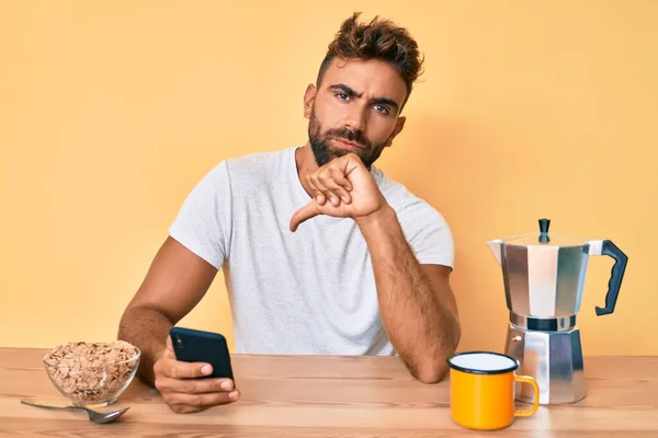Young hispanic man sitting on the table having breakfast and using smartphone with angry face, negative sign showing dislike with thumbs down, rejection concept