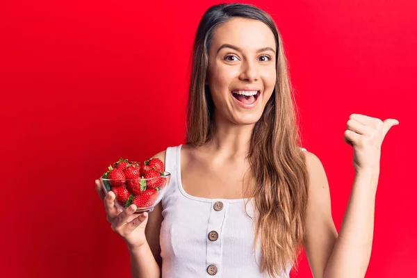 Joven Hermosa Chica Sosteniendo Tazón Con Fresas Señalando Pulgar Hacia —  Fotos de Stock