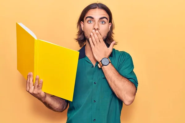 Joven Hombre Guapo Sosteniendo Libro Cubriendo Boca Con Mano Sorprendido — Foto de Stock