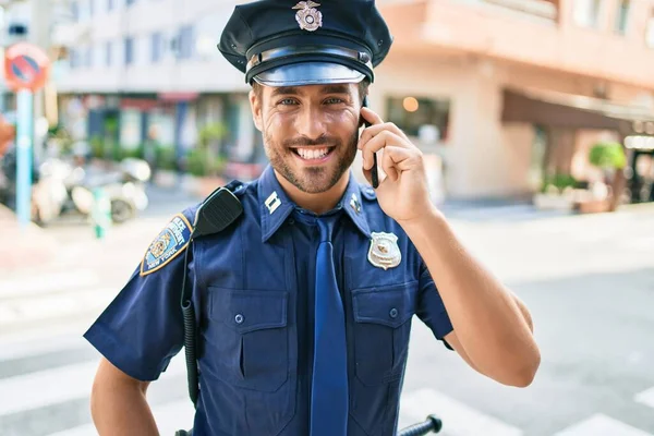 Genç, yakışıklı İspanyol polis memuru polis üniforması giyip mutlu gülümsüyor. Yüzünde tebessümle dikilip şehrin caddesinde akıllı telefondan konuşuyor..