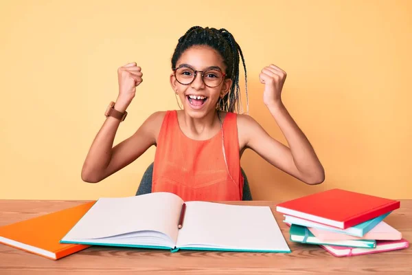 Young african american girl child with braids studying for school exam screaming proud, celebrating victory and success very excited with raised arms