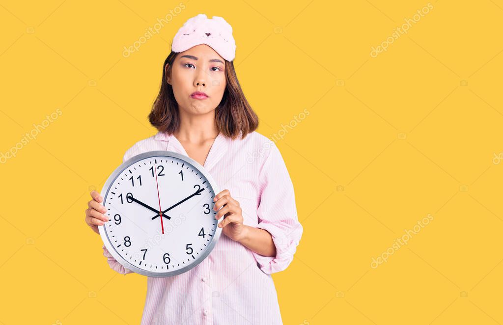 Young beautiful chinese girl wearing sleep mask and pajama holding big clock thinking attitude and sober expression looking self confident 