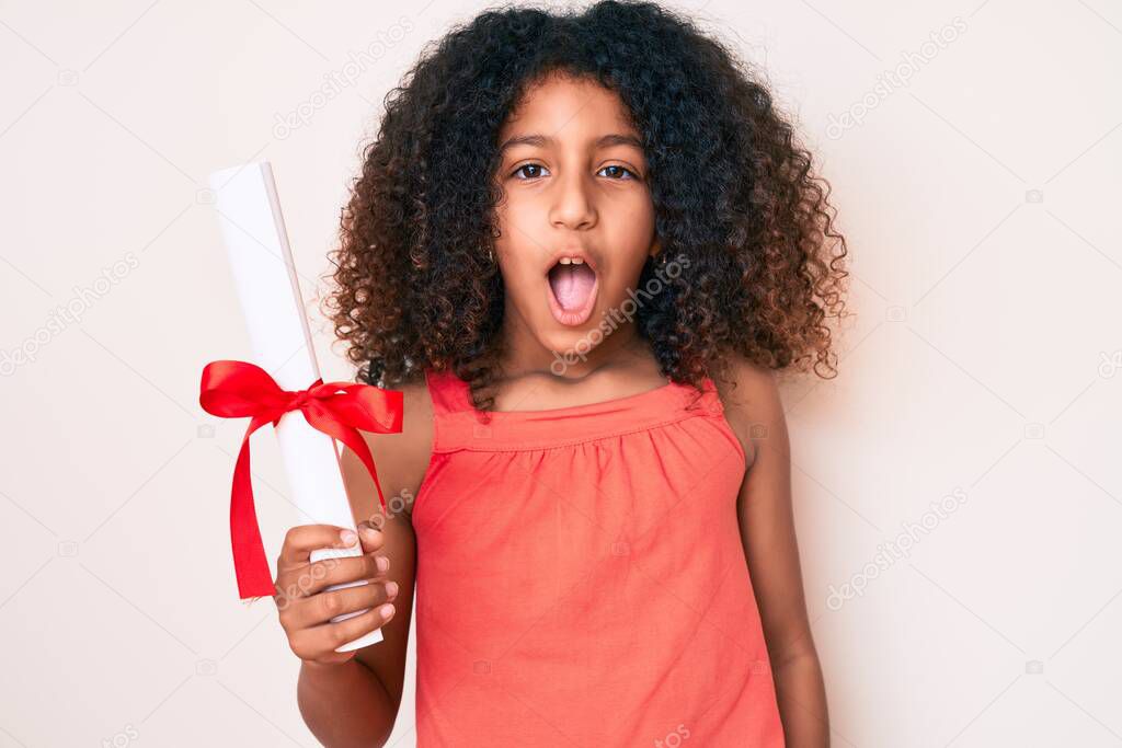 African american child with curly hair holding graduate degree diploma scared and amazed with open mouth for surprise, disbelief face 