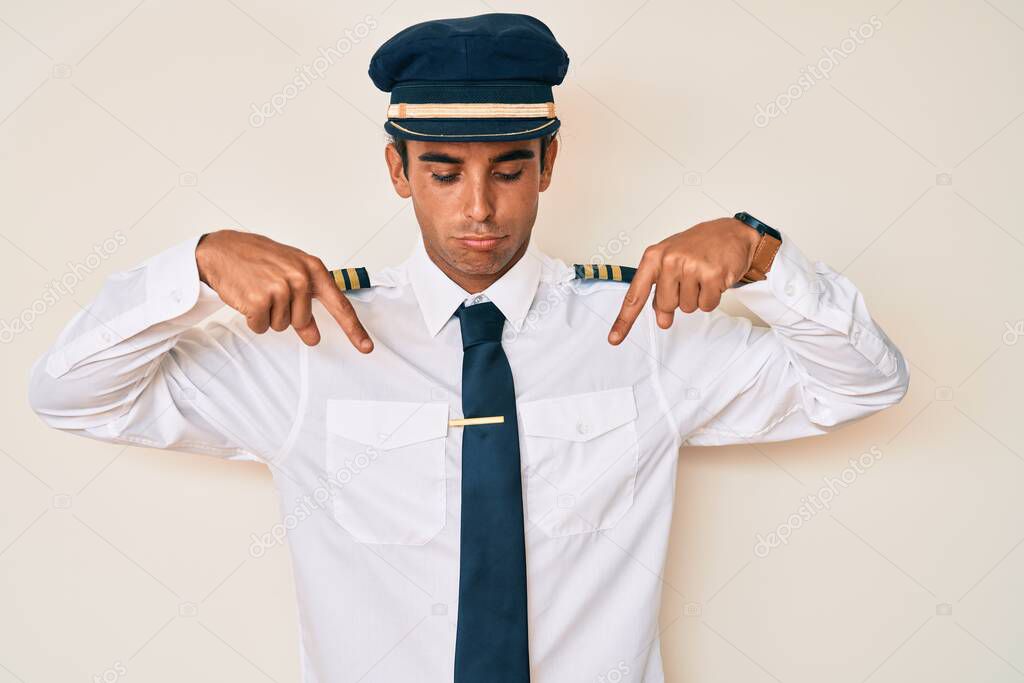 Young hispanic man wearing airplane pilot uniform pointing down looking sad and upset, indicating direction with fingers, unhappy and depressed. 