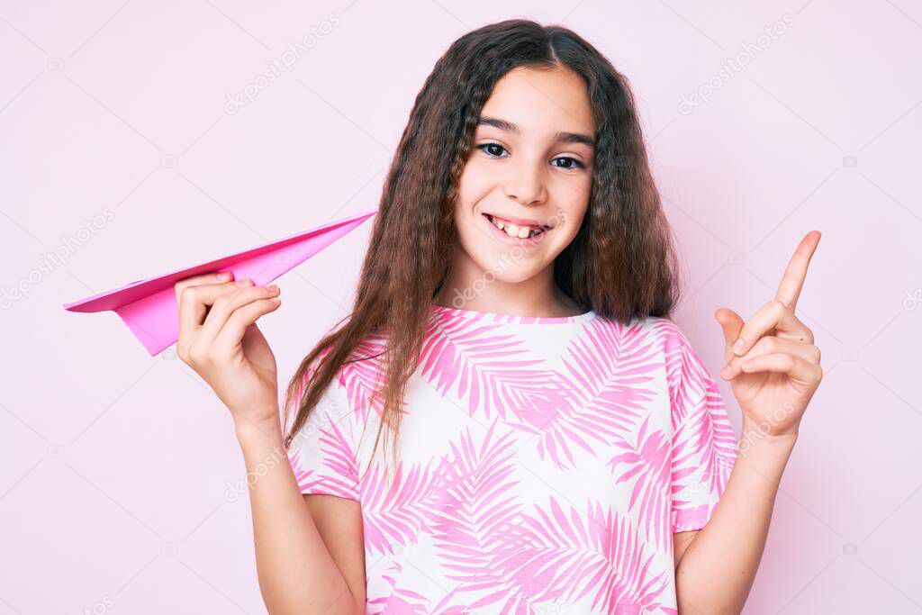 Cute hispanic child girl holding paper airplane smiling happy pointing with hand and finger to the side 