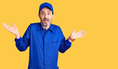 Orta yaşlı, tamirci üniforması giyen, kafası karışmış, elleri ve kolları havada bir adam. şüphe kavramı. 