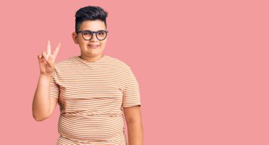 Gündelik kıyafetler giyen ve gözlüklü küçük çocuk iki numarayı gösterip işaret ederken kendinden emin ve mutlu bir şekilde gülümsüyor.. 