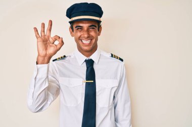 Uçak pilotu üniforması giyen genç İspanyol adam gülümsüyor ve el ve parmaklarıyla 