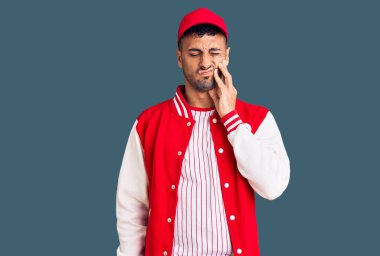 Beyzbol üniforması giyen genç İspanyol bir adam diş ağrısı ya da diş ağrısı yüzünden ağızlarına acı dolu bir ifadeyle dokunuyor. dişçi 