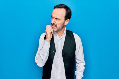 Orta yaşlı yakışıklı bir adam zarif gömlek ve yelek giyiyor. Mavi arka plan üzerinde. Şüphe üzerine yoğunlaşmış.