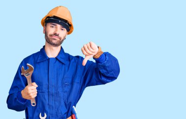 Elektrikçi üniforması giyen genç İspanyol adam elinde kızgın yüzlü İngiliz anahtarı, olumsuz işaret baş parmakları aşağıda, reddedilme konsepti. 