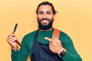 Önlük takan genç Arap adam berber jiletini tutarken gülümsüyor mutlu bir şekilde eli ve parmağı işaret ediyor. 