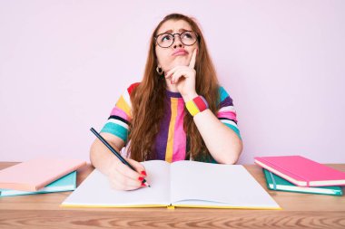 Genç kızıl saçlı kadın masada oturmuş kitap yazıyor ciddi bir yüz ifadesi var eli çenesinde sorular soruyor, kafa karıştırıcı bir fikir hakkında düşünceli. 