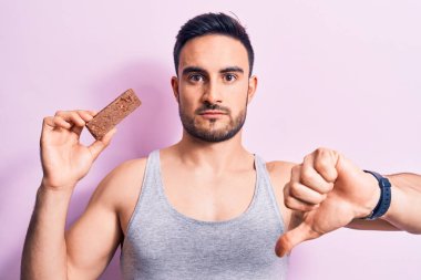 Sakallı, enerji proteini yiyen genç yakışıklı adam izole pembe arka planda kızgın yüzlü, negatif işaretli, başparmakları aşağıda, reddedilme kavramından hoşlanmadığını gösteren.