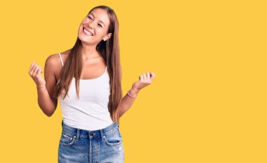 Genç ve güzel İspanyol bir kadın gündelik kıyafetler giyip çok mutlu ve heyecanlı bir şekilde kolları havada, gülümseyen ve başarı için bağıran kazanan bir jest yapıyor. kutlama konsepti. 