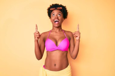 Genç Afro-Amerikalı kadın bikini giyip şaşkın şaşkın yukarı bakıyor ve parmaklarıyla işaret ediyor ve kollarını kaldırıyor.. 