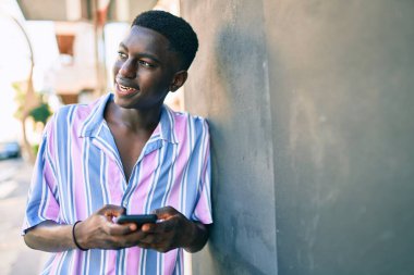 Afro-Amerikalı genç adam mutlu gülümsüyor. Şehrin caddesinde duvara yaslanmış akıllı telefon kullanıyor..