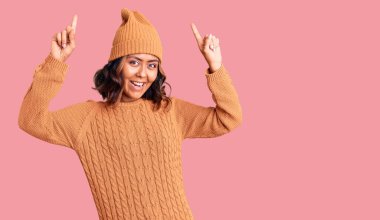 Yün kazak ve kış şapkası giyen genç, güzel, melez bir kadın. Şaşırmış, şaşırmış ve parmaklarıyla işaret ediyor ve kollarını kaldırıyordu.. 