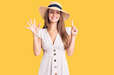 Genç, sarışın, yaz elbisesi ve şapkası giyen, altı numaralı parmaklarıyla işaret eden, kendinden emin ve mutlu bir şekilde gülümseyen bir kadın.. 